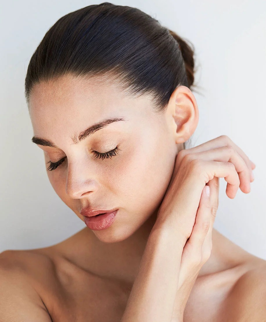 Close-up of a woman with light-toned skin and dark hair in a bun. Her eyes are closed, and her expression is serene. Her hands rest gently on her neck and face. The background is a plain light gray or white, emphasizing her peaceful features. - Facetite in Owings Mills, MD
