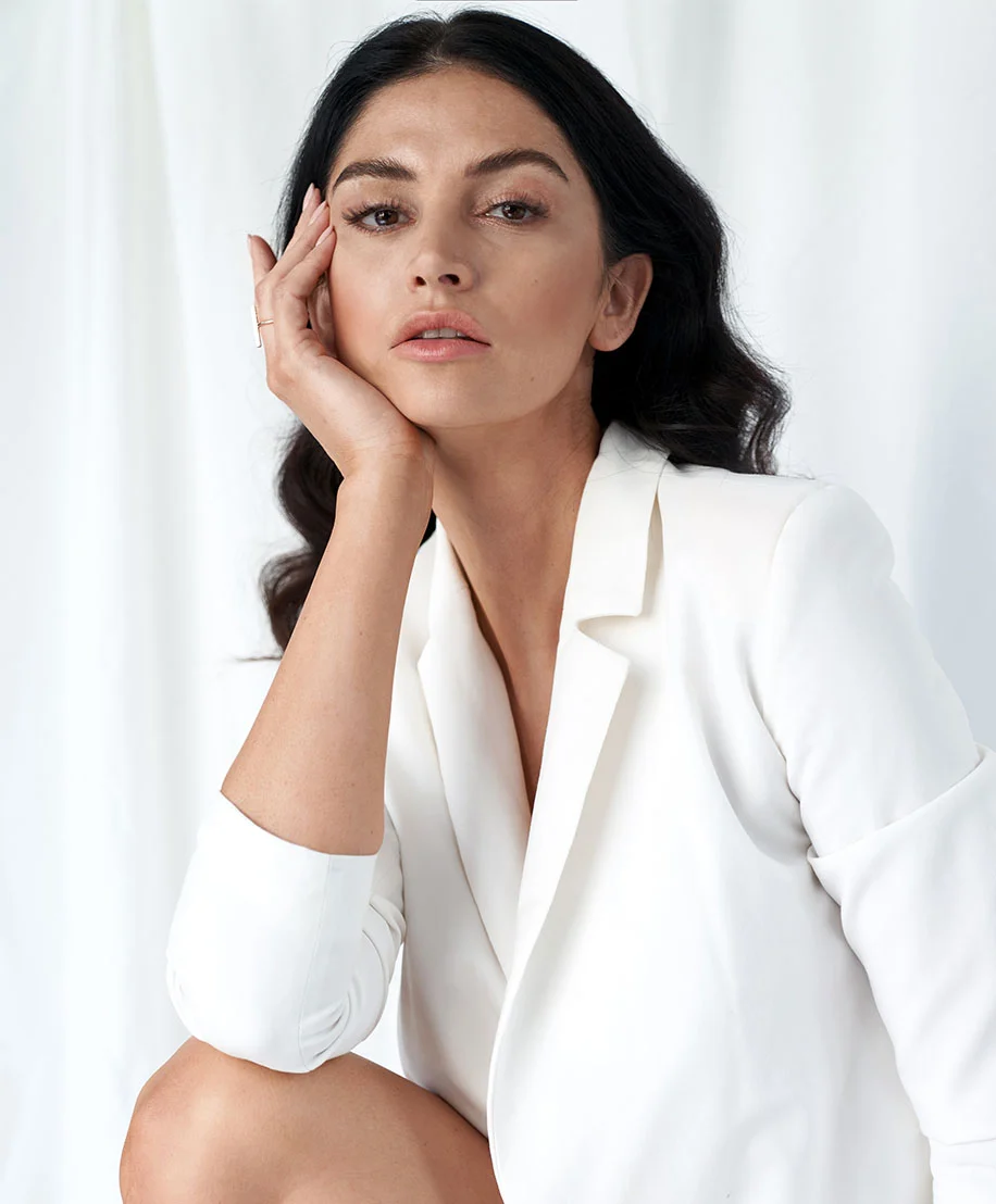 A woman with long dark hair sits wearing a white blazer with a notched lapel. Her head is slightly tilted, and her hand rests on her cheek. She has a neutral expression, light to medium tan skin, and is looking directly at the camera. A simple ring is visible on one finger. The background is plain off-white with soft lighting highlighting her features. - Morephus8 in Owings Mills, MD