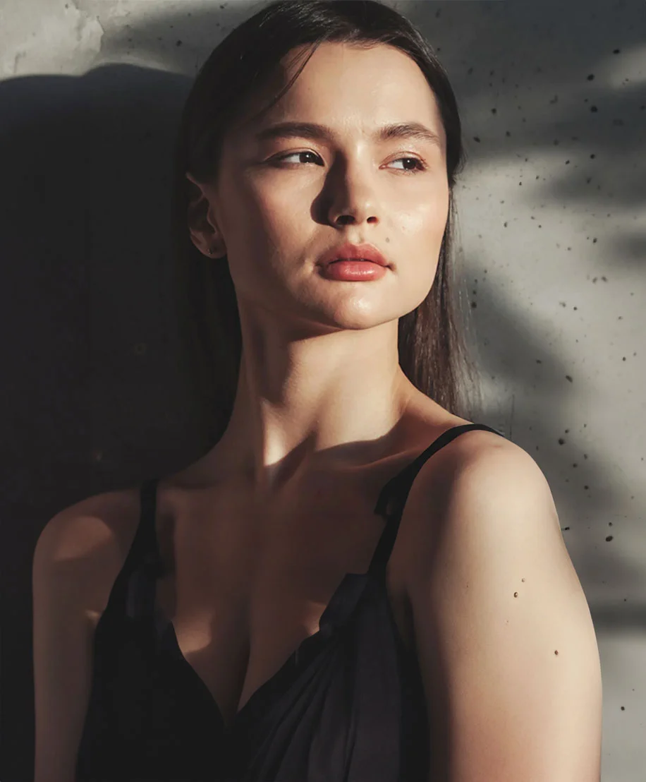 Young woman with long dark hair stands against a textured gray concrete wall, wearing a black sleeveless top. She is angled slightly toward the camera with a neutral expression. Sunlight casts a face-shaped shadow on the wall beside her, and her smooth, light-toned skin is softly highlighted. - Skin Pen in Owings Mills, MD