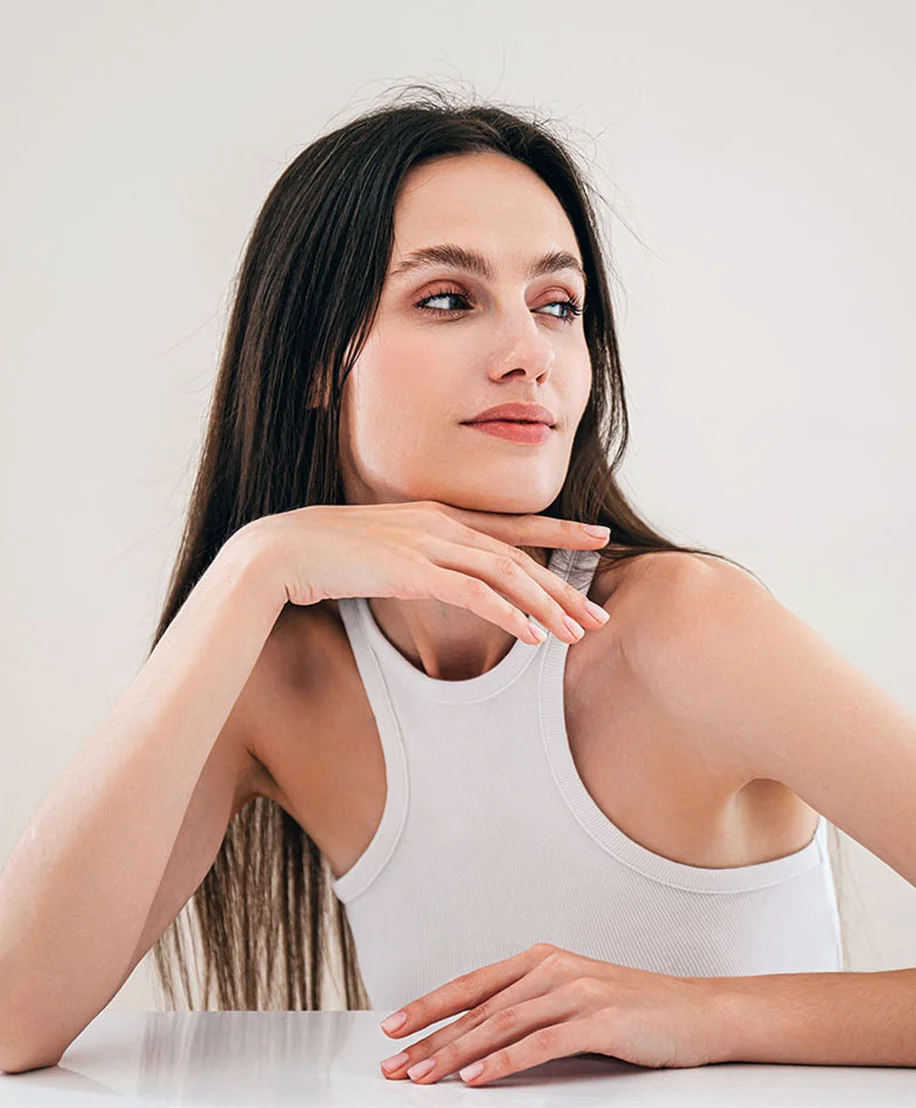 A woman with long, dark brown hair sits with her head turned slightly to the right, resting her chin on her hand. She wears a simple white tank top and has smooth, light-toned skin. Her gaze is directed off-frame, and the background is a pale cream color. Soft lighting highlights her natural features and the graceful position of her hands and arms. - ZO Skinhealth in Owings Mills, MD